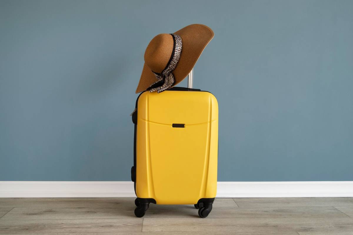  A bright yellow suitcase with black wheels sits against a blue wall, topped with a stylish brown sunhat.