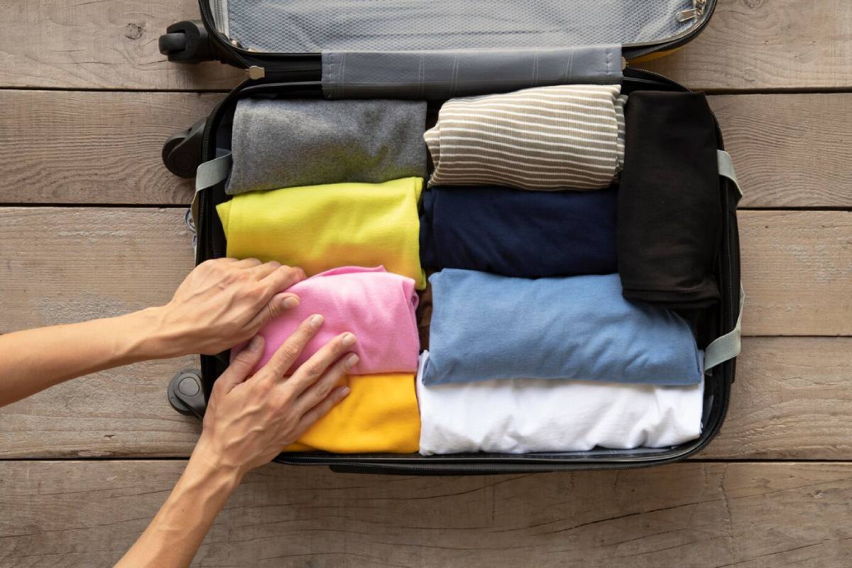 A neatly packed suitcase filled with colorful rolled t-shirts, with hands arranging the items, on a rustic wooden surface. 