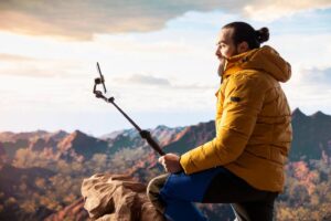 A person in a yellow jacket sits on a rock, holding a selfie stick, overlooking a scenic mountainous landscape at sunset.