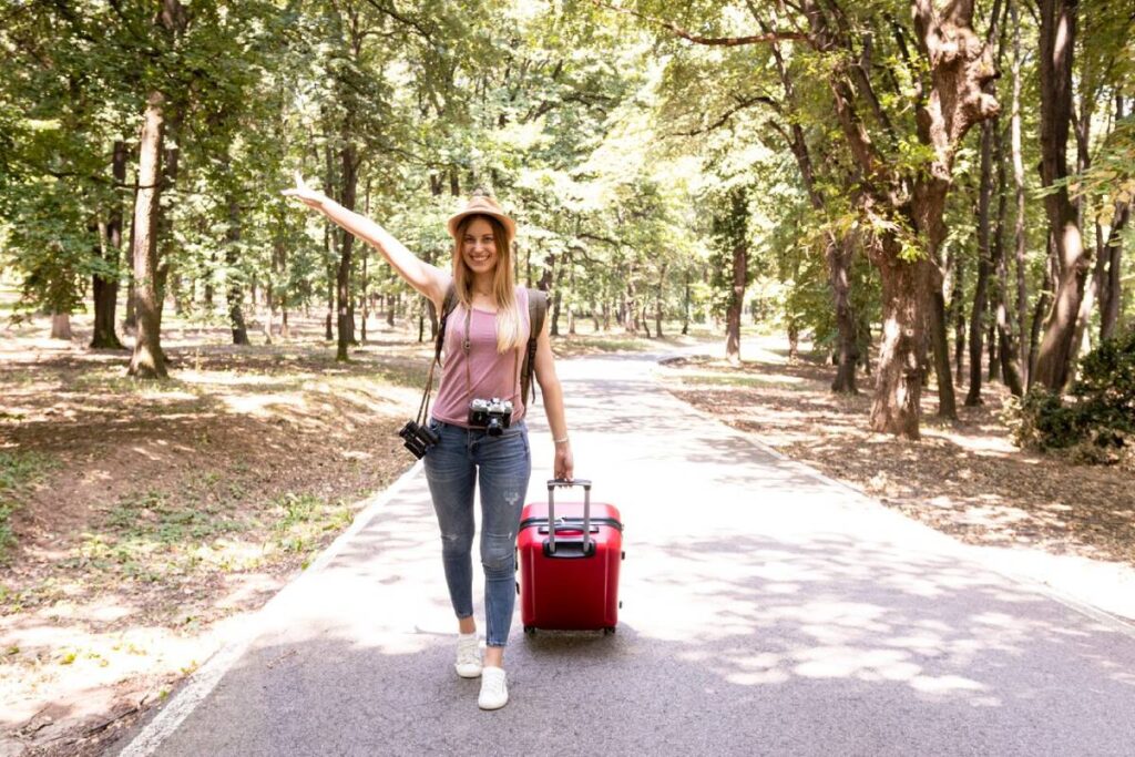 A young woman stands in a forested area, waving while pulling a red suitcase, wearing a hat and a camera around her neck.
