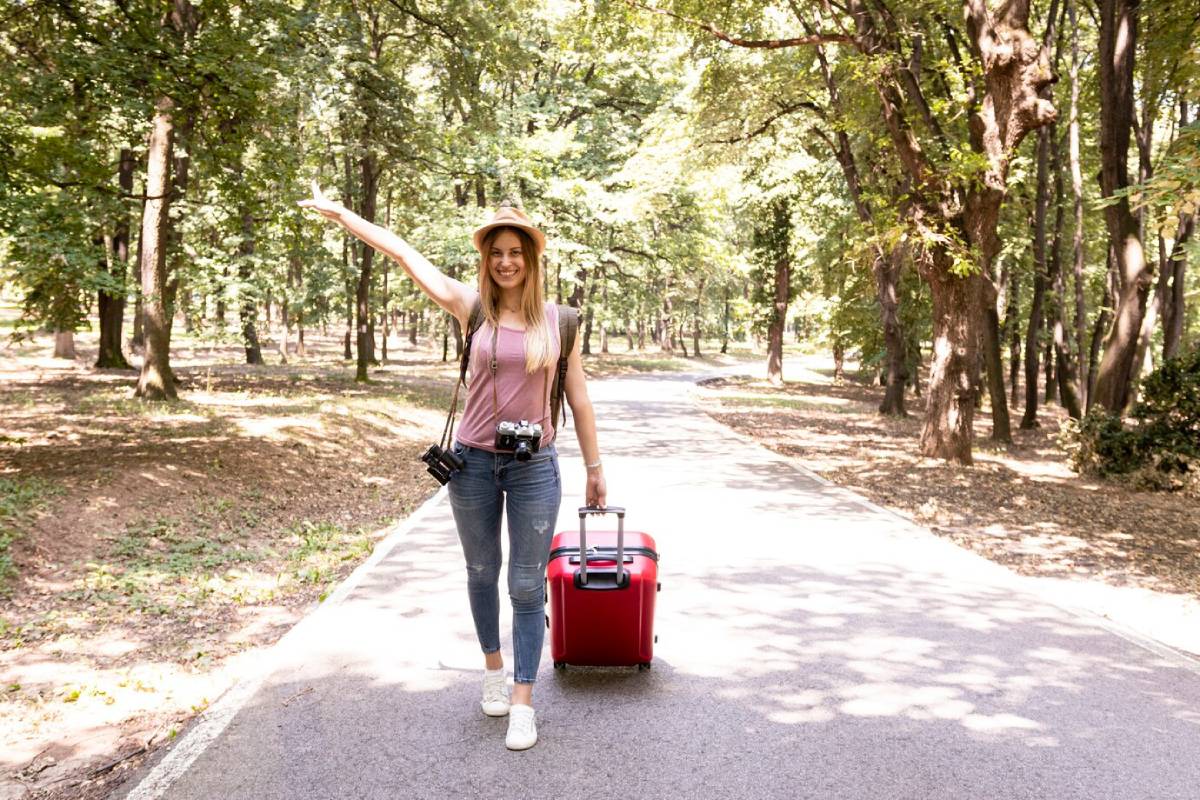 A young woman stands in a forested area, waving while pulling a red suitcase, wearing a hat and a camera around her neck.