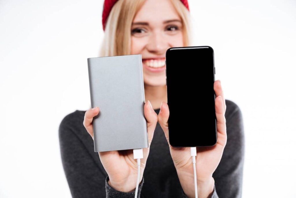 A person holds a silver power bank in one hand and a black smartphone with a charging cable in the other, set against a white background.