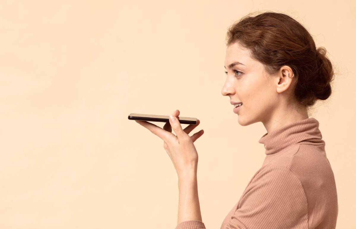 A person in a beige turtleneck sweater holds a smartphone to their ear against a soft pastel background. 