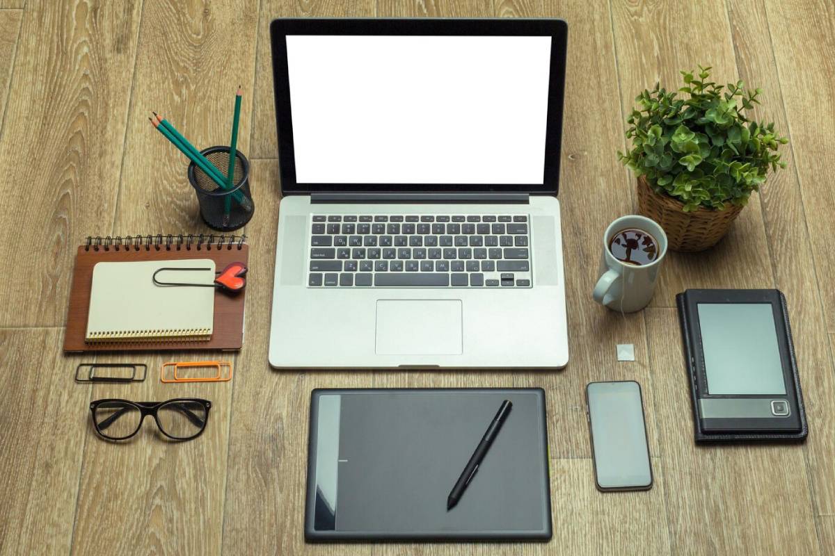 A workspace featuring a laptop, tablet, notebook, glasses, stationery, a coffee mug, and a small plant, arranged on wooden flooring. 