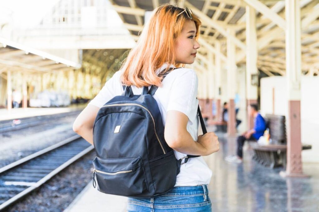 A person stands with a black backpack in a bright train station, surrounded by railway tracks and benches, ready for travel.