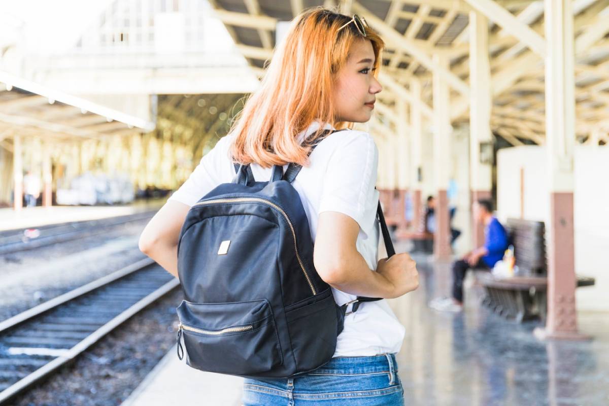 A person stands with a black backpack in a bright train station, surrounded by railway tracks and benches, ready for travel.