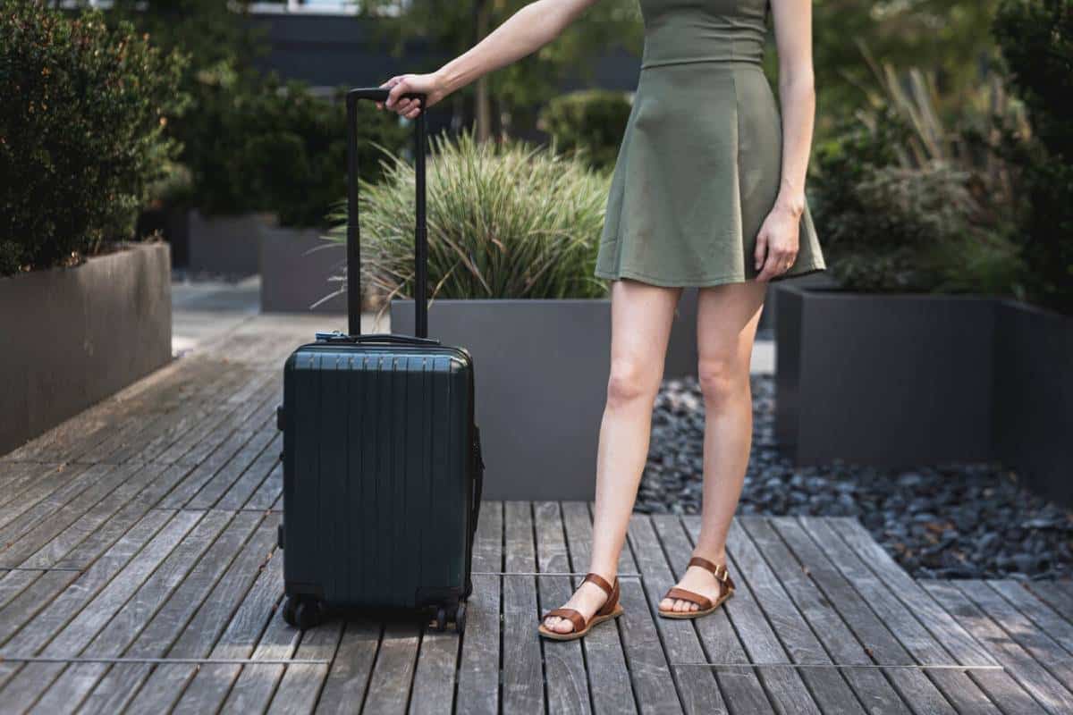 A woman in a green dress stands beside a dark suitcase on a wooden pathway, surrounded by neatly trimmed plants.
